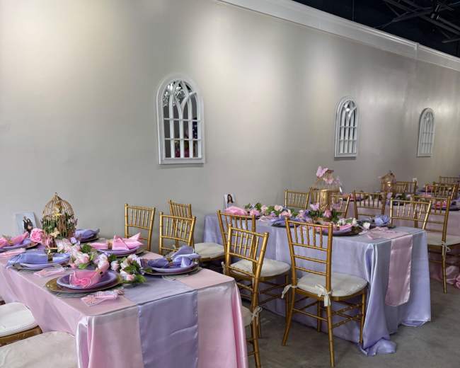 The image shows a dining area with several tables covered in pink and purple satin tablecloths, set with plates and decorative centerpieces.