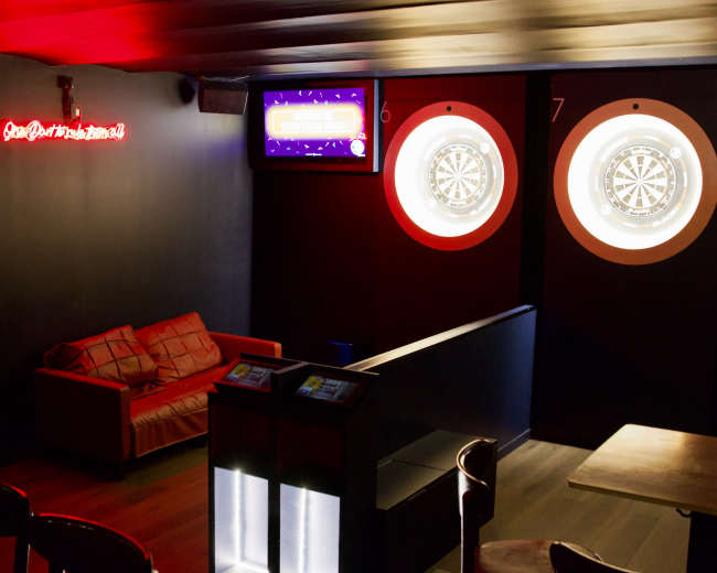 The image shows a dimly lit bar area featuring a red neon sign, a leather couch, and two dartboards illuminated on the wall.