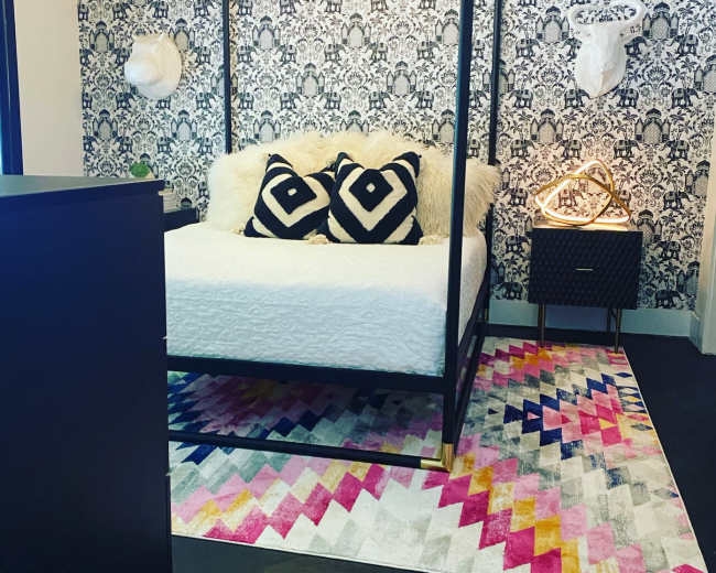The image shows a modern bedroom featuring a canopy bed with a white bedspread, black and white patterned throw pillows, decorative wall art, and a colorful geometric rug.