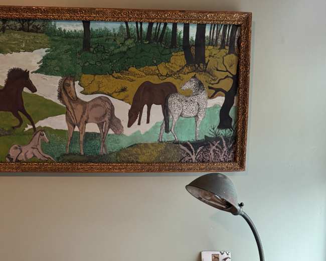 A wooden desk with an open notebook, a lamp, and a decorative panel of horses is located against a green wall.