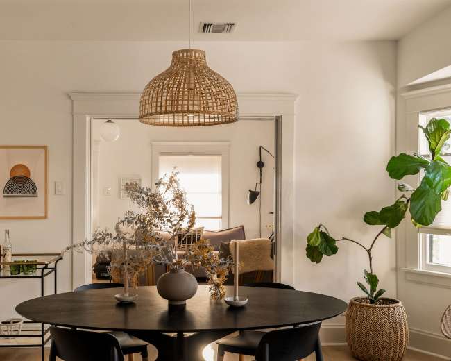 A round black dining table is set in a well-lit room featuring a rattan pendant light overhead and a decorative arrangement of dried branches.