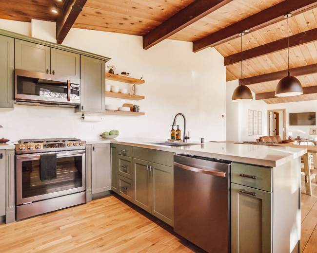 A modern kitchen features green cabinetry, stainless steel appliances, and a large island with a seamless countertop, all under a wooden beamed ceiling.
