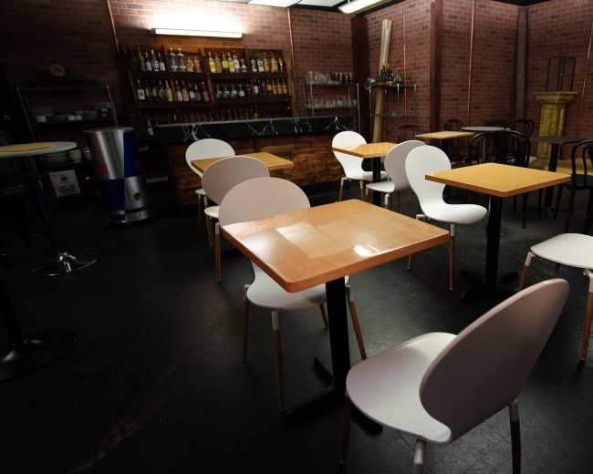 The image shows an empty restaurant interior with several tables and chairs arranged on a dark floor, surrounded by brick walls and a bar area in the background.