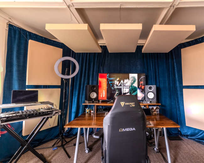 A music production setup featuring a desk with a keyboard, speakers, and a large monitor surrounded by acoustic panels and blue curtains.