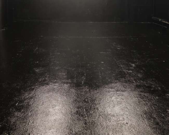 The image shows a large, empty black room with polished flooring and overhead lighting.