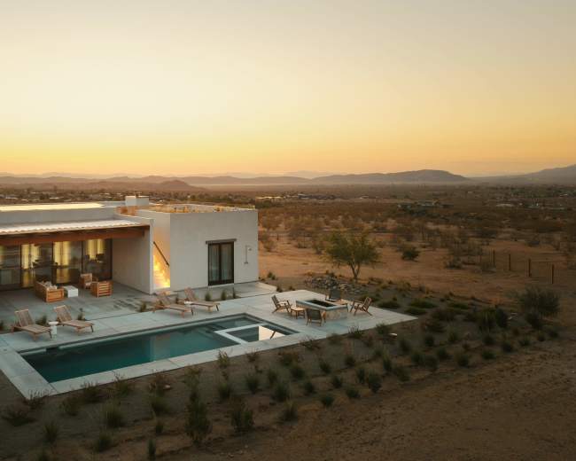 A modern house with a pool sits among desert landscaping, framed by a sunset over distant mountains.