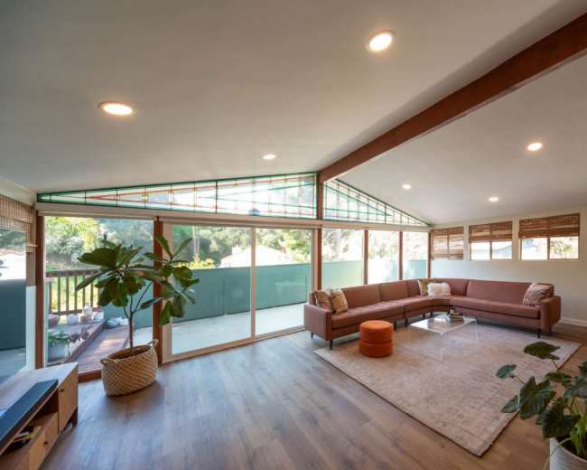 The image shows a spacious living room featuring a brown sectional sofa, a glass coffee table, hardwood flooring, and large windows letting in natural light.