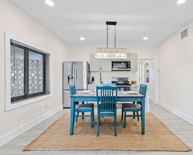 The image shows a modern kitchen with a dining table and chairs, stainless steel appliances, and a large window, all set over a woven rug.