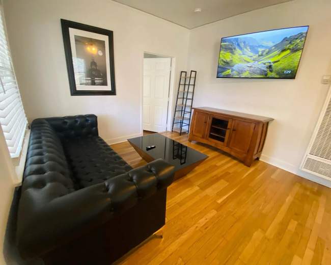 The image shows a living room with a black tufted couch, a glass coffee table, a wooden media console, and a wall-mounted television displaying a landscape.