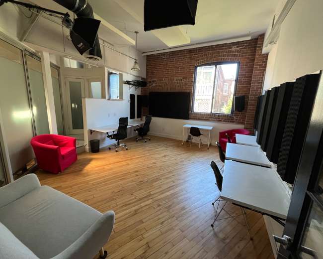 The image shows a spacious office area with wooden floors, a large window, a whiteboard, and various furniture including chairs and desks.