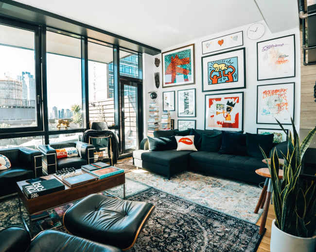 The image shows a modern living room with large windows, a black sectional sofa, various framed artwork on the walls, and a wooden coffee table on a patterned rug.