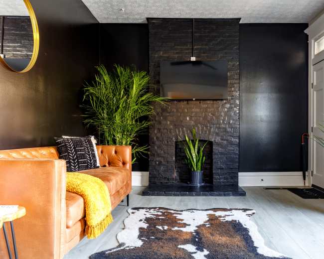 The image shows a modern living room featuring a brown leather sofa, a black stone fireplace with a mounted television, and green plants beside the fireplace and entrance.