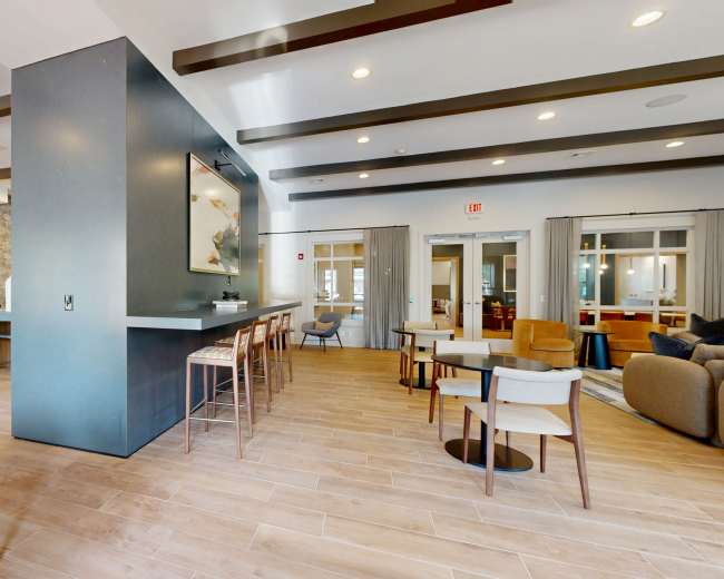The image shows a modern interior of a community space featuring a mix of seating options, a bar area, and natural lighting.