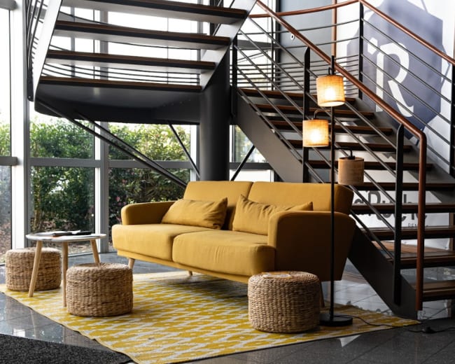 A yellow sofa is placed next to a table and two woven stools on a patterned rug in a space with large windows and a spiral staircase.