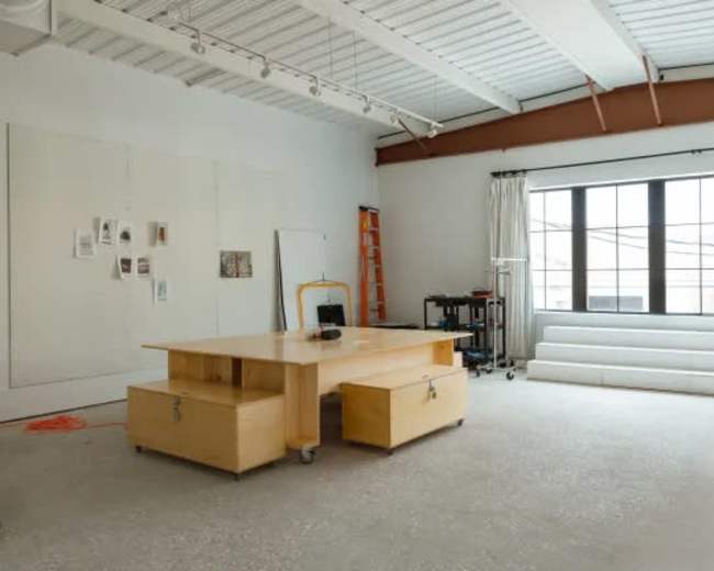 The image shows a spacious, minimalist office or studio featuring a central wooden desk, a few pinned documents on the wall, a ladder, and white furniture arranged by a large window.