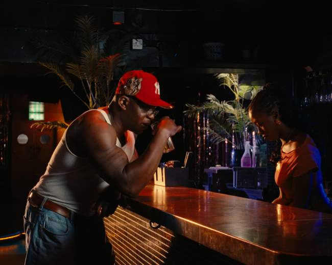 A man in a tank top and baseball cap leans on a bar while a woman stands behind it.