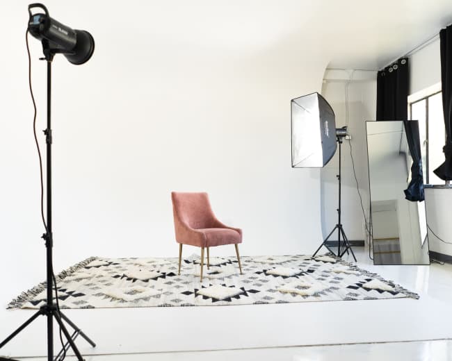 The image shows a minimalistic photography studio featuring a pink chair on a patterned rug, with lighting equipment and a large mirror against a plain white backdrop.