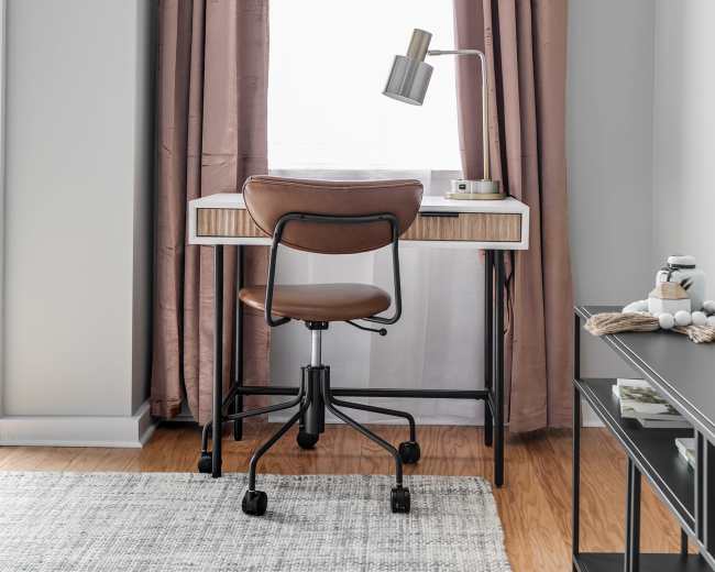 A home workspace features a desk with a chair, a lamp, and a large window dressed in brown curtains.