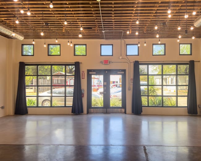 The image shows a spacious, well-lit interior with large windows and an exit door, featuring string lights overhead and a polished concrete floor.