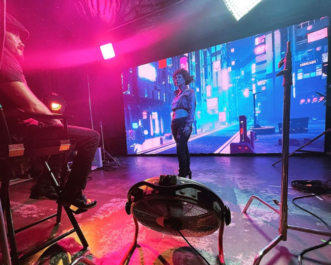 A person sits in a director's chair in front of a large screen displaying a vibrant cityscape, while another person stands on a set illuminated by colorful lights.
