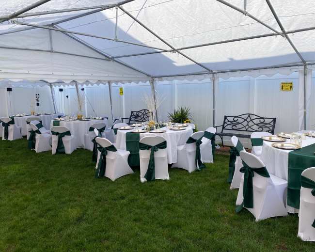 A tent is set up on grass, featuring several tables with white tablecloths and green sashes, arranged for a festive gathering.