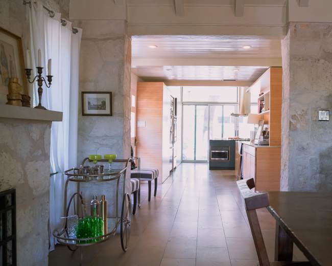 The image shows a modern interior space with a kitchen visible at the end, flanked by a dining area and a cart with glassware in the foreground.
