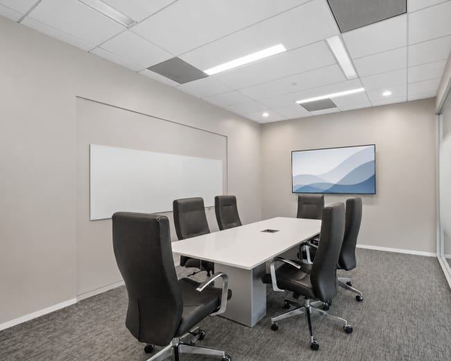 A modern conference room features a long white table surrounded by black office chairs, with a large screen mounted on the wall and glass panels on one side.