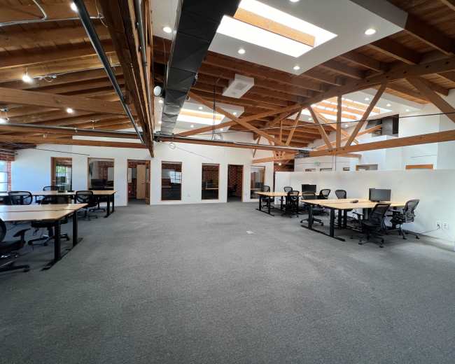 The image shows a spacious office interior with wooden beams, several desks arranged in clusters, and large windows allowing natural light to enter.