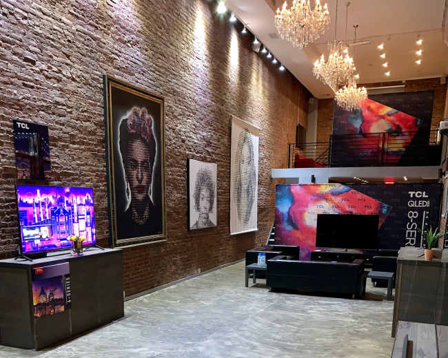 The image shows a modern showroom with a polished concrete floor, featuring various large artworks on the brick walls and multiple television displays.