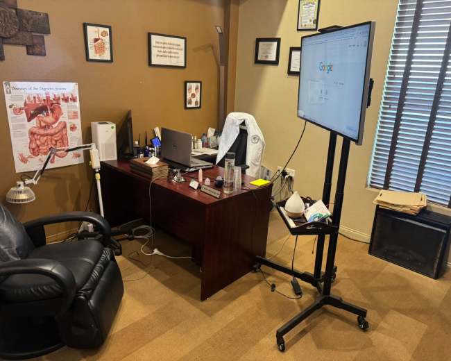 The image shows a medical office with a dark wooden desk, a computer monitor on a stand, medical textbooks, and a white coat hanging nearby.