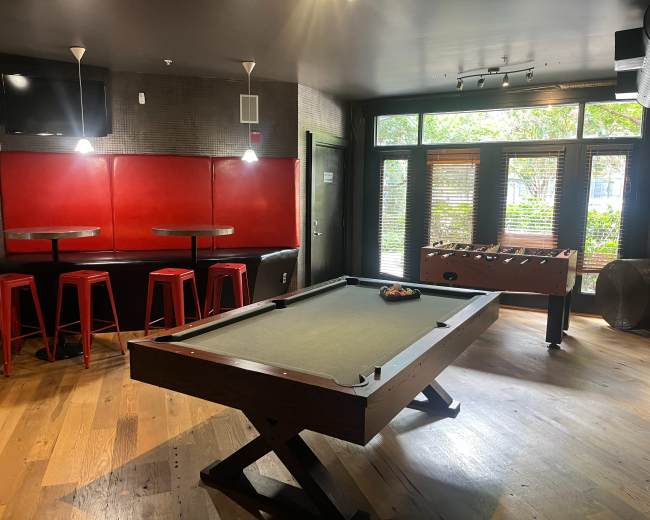 The image shows a game room with a pool table, foosball table, and bar seating against red walls with large windows letting in natural light.
