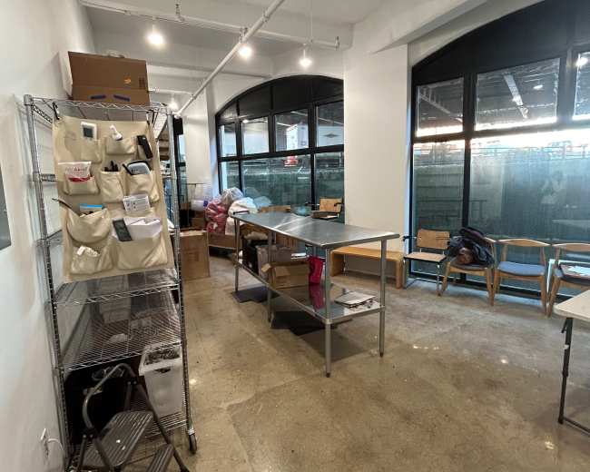 The image shows a large, empty room with a concrete floor, tables in the center, shelves with storage bags on one side, and large windows letting in natural light.