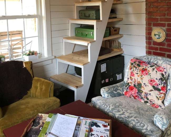 The image shows a cozy interior space with a white wooden staircase leading to an upper level, surrounded by colorful furniture and shelves.
