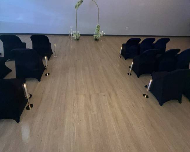 The image shows a ceremony setup with rows of dark chairs facing an arch decorated with flowers, illuminated by string lights overhead.