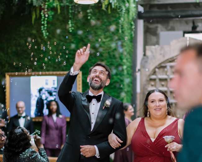 A man in a tuxedo and a woman in a red gown walk together while bubbles float around them in a decorated indoor space.