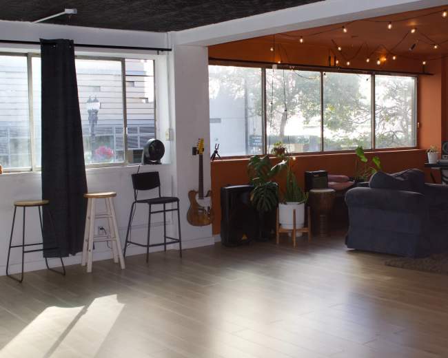The image shows a spacious room with hardwood flooring, large windows lined with string lights, and various furnishings, including stools, a couch, and a music setup.