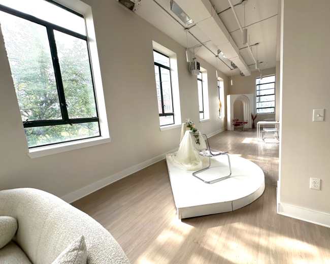 The image shows a spacious, modern interior with large windows, showcasing a small stage with a decorative drape and a chair amidst clean, minimalist decor.