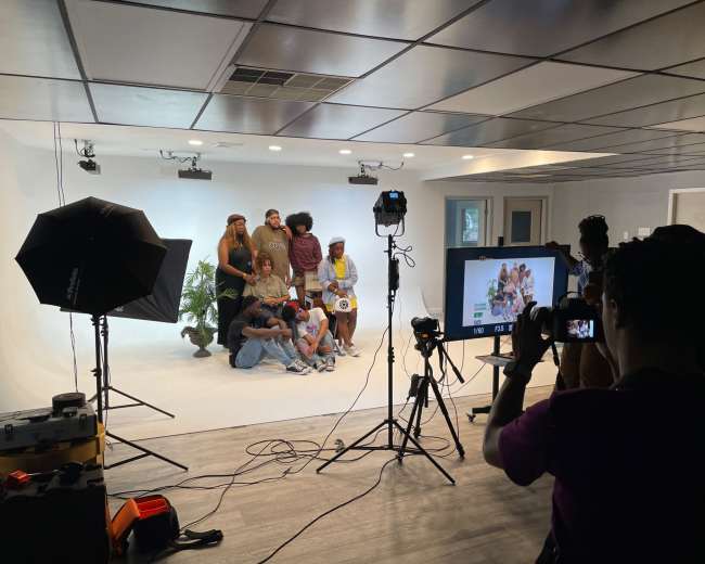 A group of people poses for a photo in a studio with a white backdrop, while others set up cameras and lighting equipment.