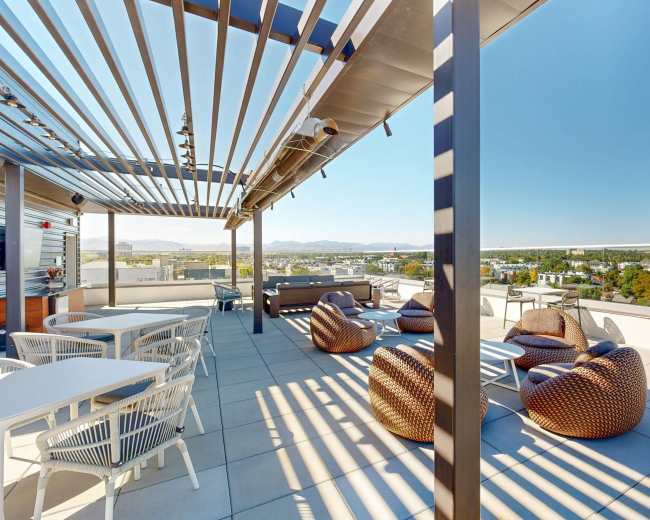 The image shows a rooftop terrace featuring lounge chairs, tables, and a panoramic view of the surrounding landscape under a clear blue sky.