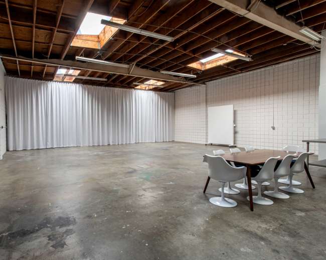 The image shows a spacious, minimalist meeting room with a wooden table and six white chairs, set against a backdrop of plain walls and a ceiling with exposed beams.