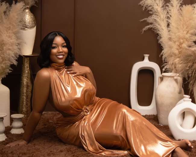 A person in a metallic copper dress is reclining on a brown rug surrounded by decorative vases and dried plants against a brown backdrop.