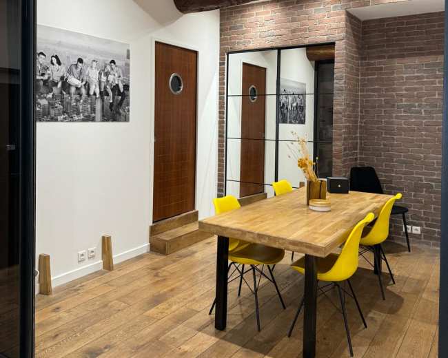 A modern conference room features a wooden table surrounded by yellow chairs, with exposed brick walls and large windows.