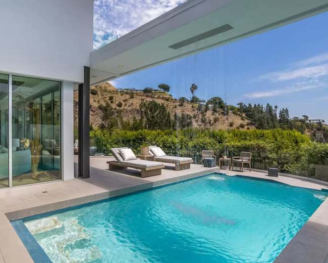 The image shows a modern outdoor pool area with lounge chairs, surrounded by greenery and a view of a hillside.