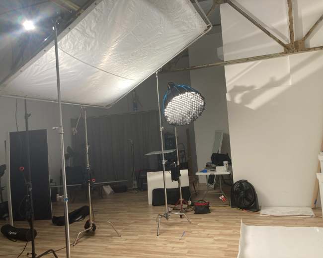 The image shows a photography studio set up with lighting equipment, a large softbox, and a circular light fixture in a room with wooden flooring and various studio props scattered around.