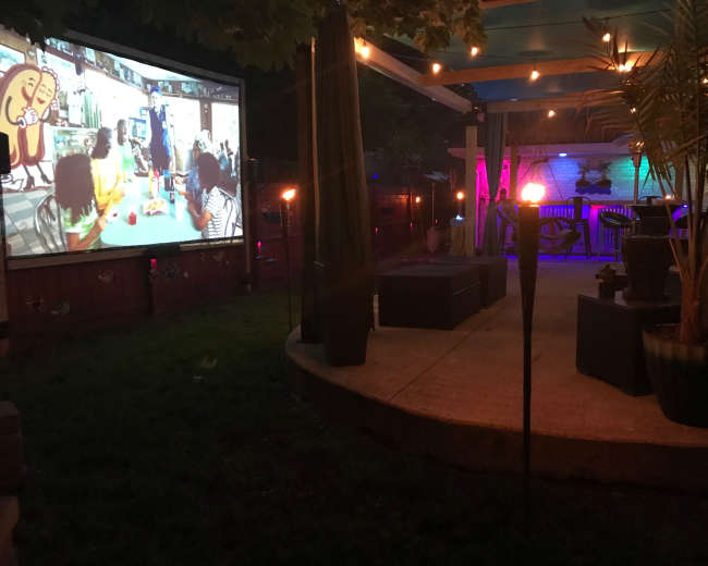 A large outdoor screen displays a colorful scene while a backyard area with seating and plants is illuminated by ambient lighting.