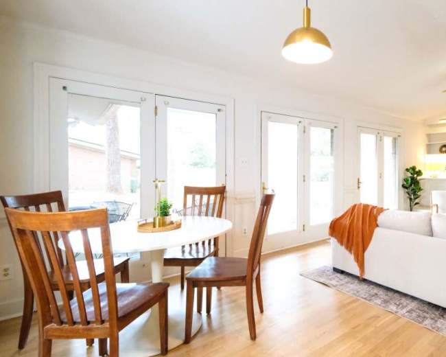 The image shows a modern living room with a light-colored couch, a circular dining table with wooden chairs, and large windows leading to an outdoor space.