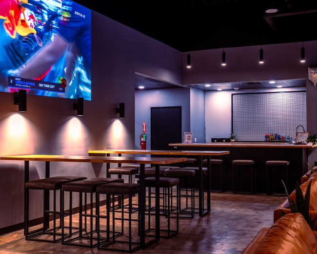 The image shows a modern bar area with high tables and stools, a large wall-mounted TV displaying sports, and a bar counter in the background.