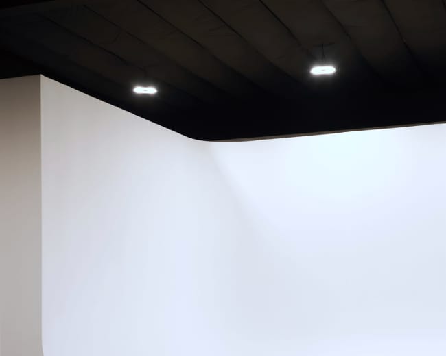 The image shows a minimalist interior space with a smooth, curved white wall and a concrete floor.