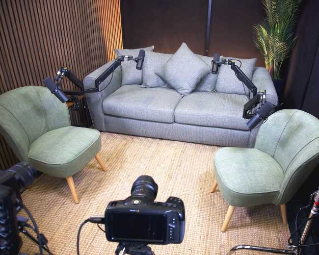 The image shows a small recording studio setup featuring a gray sofa with two green chairs positioned in front, all arranged on a jute rug, with camera equipment aimed at the seating area.