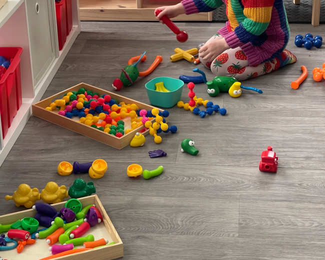 A child wearing a party hat plays with colorful toys on the floor of a playroom.